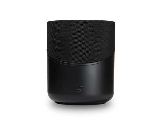 Der Bluesound PULSE M in Schwarz hat eine stoffbespannte Oberseite, eine glatte Unterseite und ein kleines Frontlogo. Dieser kabellose Lautsprecher bietet HiFi-Stereoklang und 360° Klangverteilung. Abgebildet vor einem weißen Hintergrund.