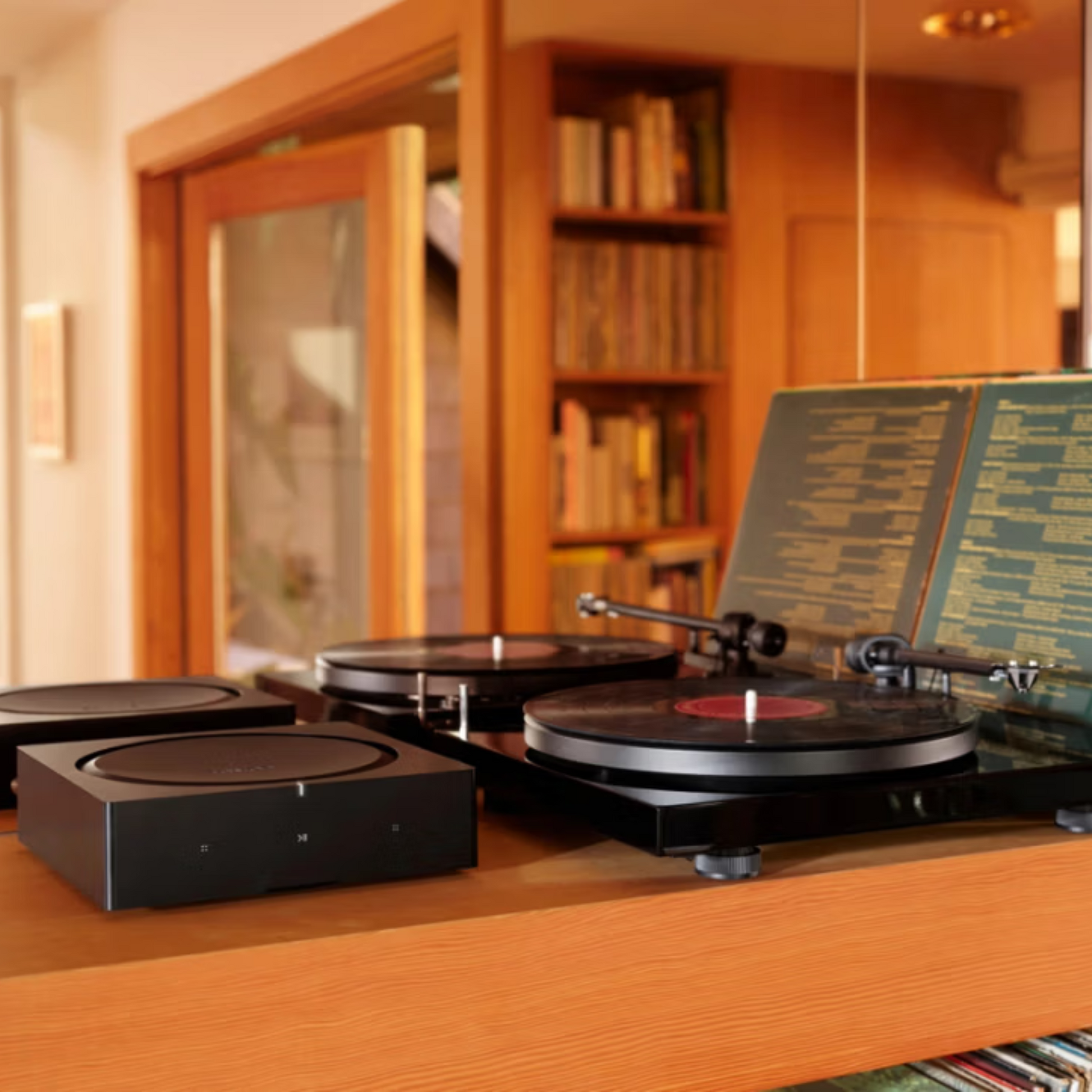 Zwei Schallplattenspieler und ein schwarzer Soundreference Sonos Amp Verstärker stehen auf einem Holzregal, umgeben von offenen Plattencovern, Bücherregalen und der einladenden Atmosphäre von Multiroom-Sound, der den Raum erfüllt.