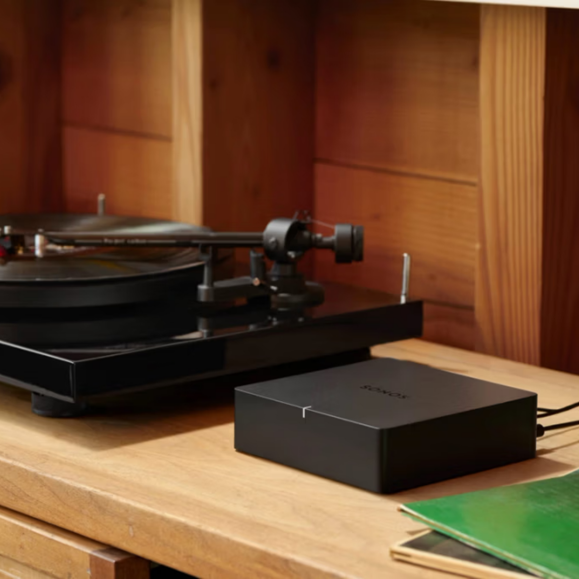Ein schwarzer Plattenspieler und der Soundreference Sonos Port Netzwerk Audio Streamer stehen auf einer Holzoberfläche in einem gemütlichen, holzgetäfelten Raum. Eine grüne Schallplattenhülle lugt hervor und beleuchtet die hochwertige Audioqualität für ein immersives Hörerlebnis.