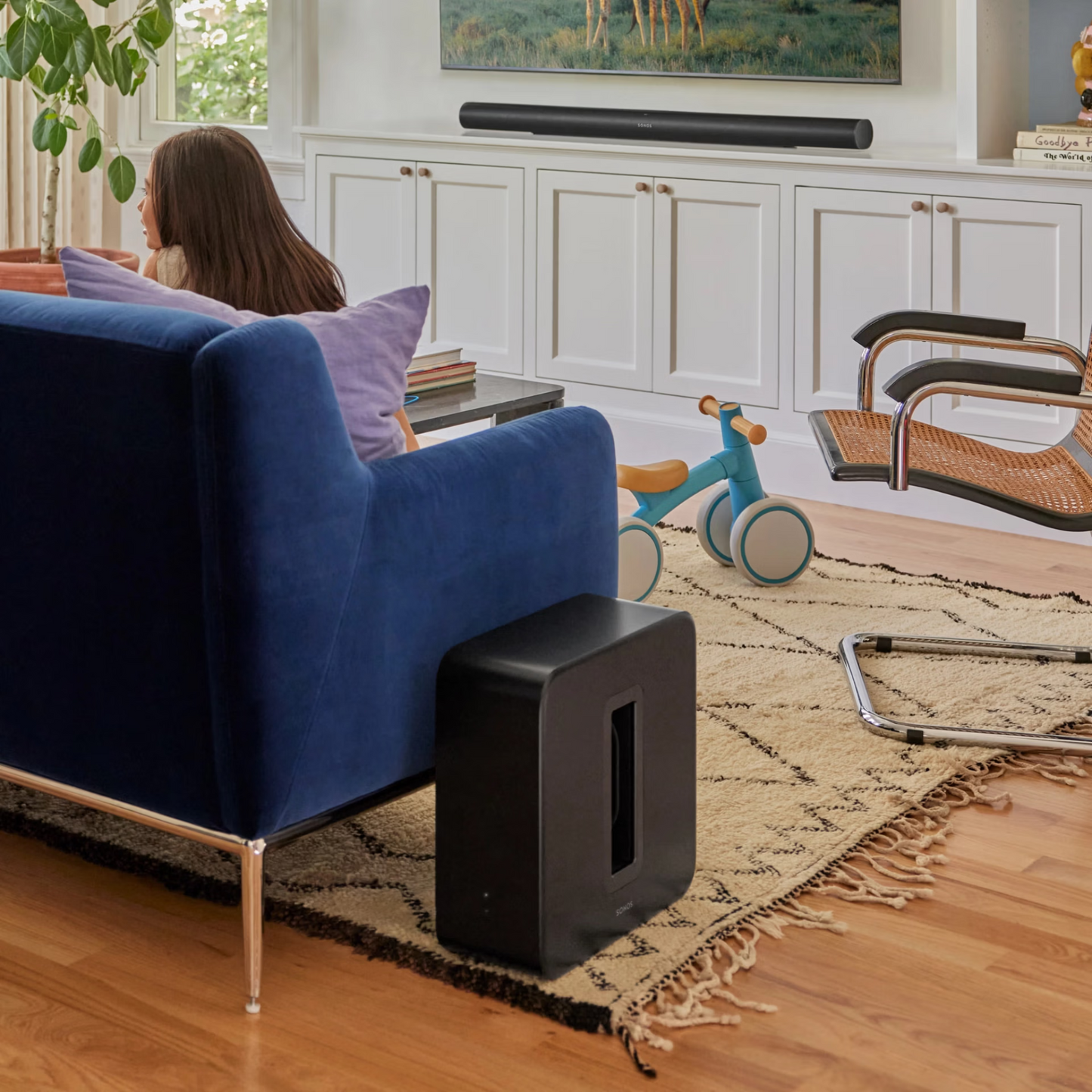 Eine Frau sitzt auf einem blauen Sofa vor einem Fernseher, während der Sonos Sub 4 Subwoofer auf einem gemusterten Teppich dynamischen Sound liefert. Ein blaues und oranges Dreirad und moderne Stühle vervollständigen das helle, ordentlich dekorierte Wohnzimmer.