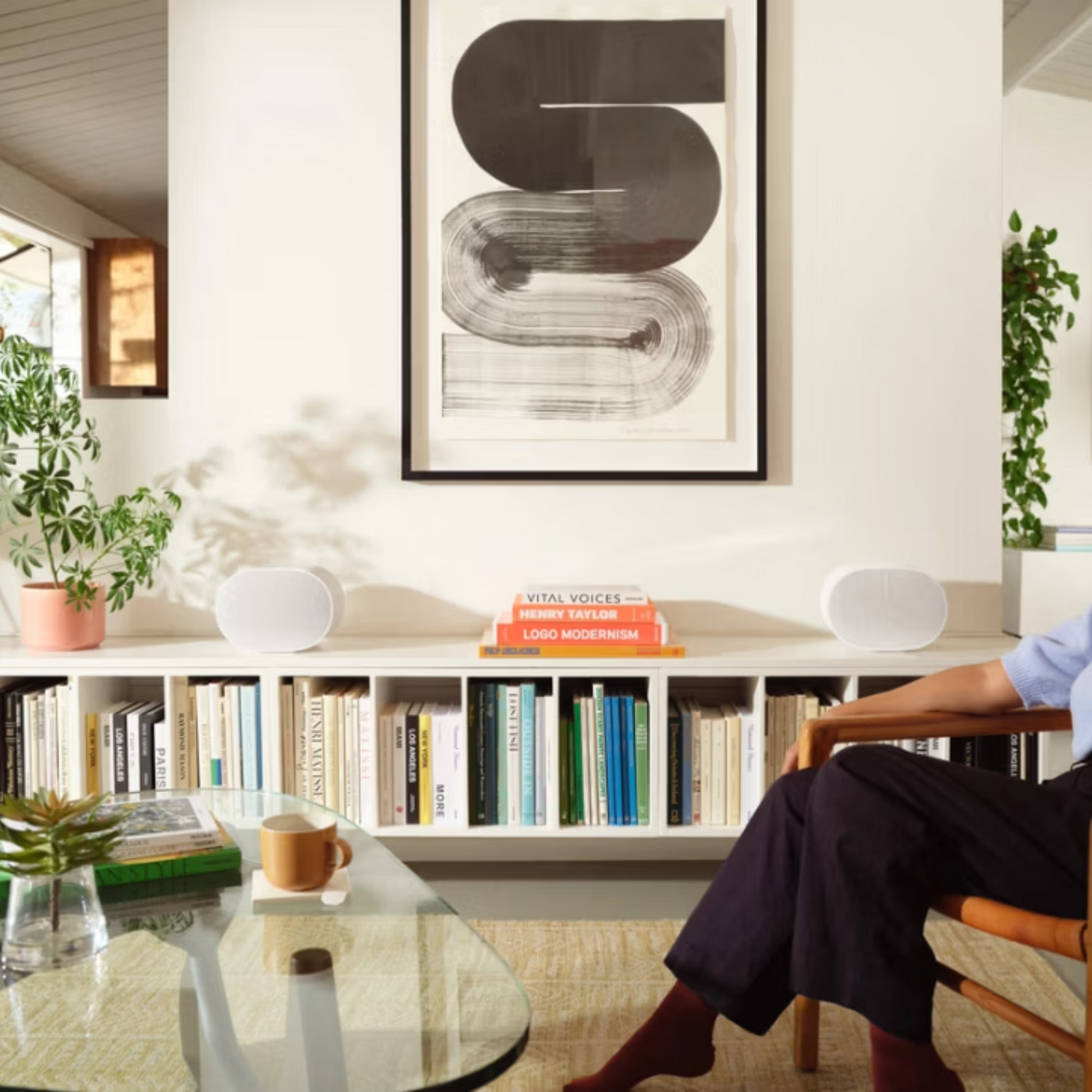 Ein modernes Wohnzimmer mit einem gläsernen Couchtisch, mit Büchern gefüllten Regalen, einer Topfpflanze, abstrakter Wandkunst und einer sitzenden Person - mit dem Sonos Era 300 Smart Speaker für immersiven Sound und eine gehobene Atmosphäre.