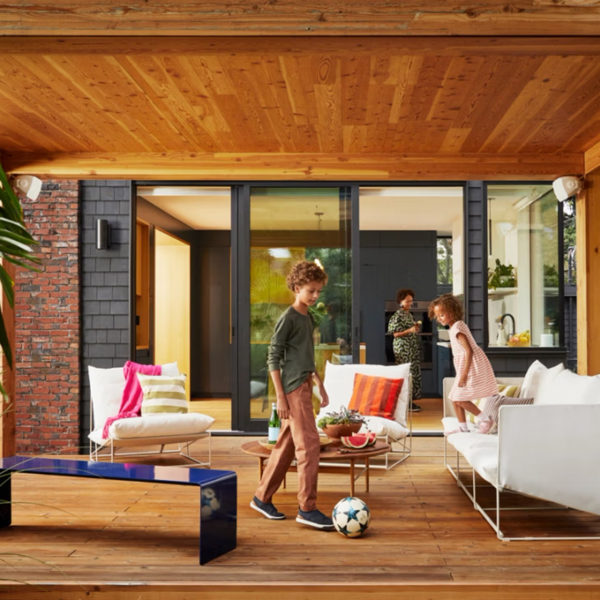 Zwei Kinder spielen auf einer Holzterrasse mit einem Fußball, während sich zwei Erwachsene in der Küche unterhalten. Große Schiebetüren verbinden die Räume, in denen die Sonos Outdoor Speaker von Sonos und Sonance Paar by Soundreference für großartigen Sound im Freien sorgen.