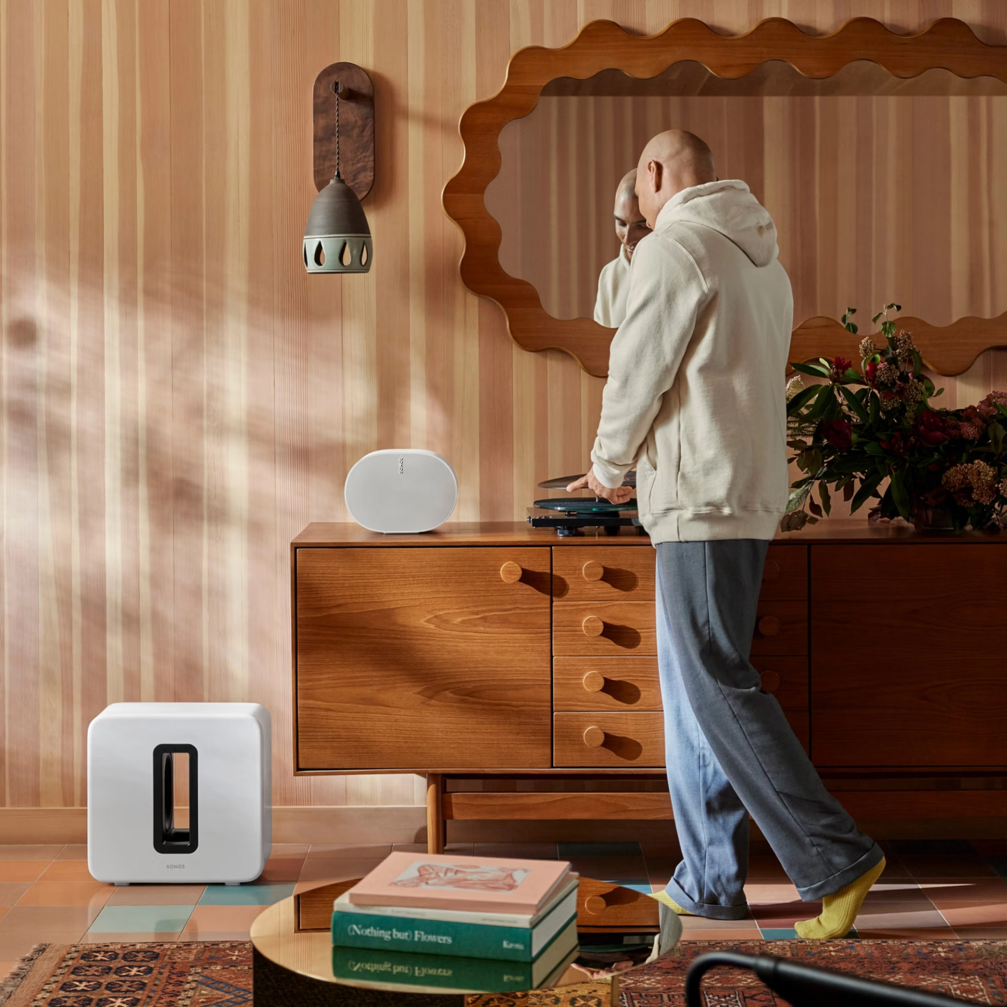 Eine Person in einem cremefarbenen Kapuzenpulli und einer blauen Hose steht neben einem hölzernen Sideboard mit einem Schallplattenspieler und dem Sonos Sub 4 Subwoofer für die dynamische Wiedergabe in einem stilvollen, im Retro-Stil gehaltenen Wohnzimmer mit gestreiften Wänden und moderner Einrichtung.