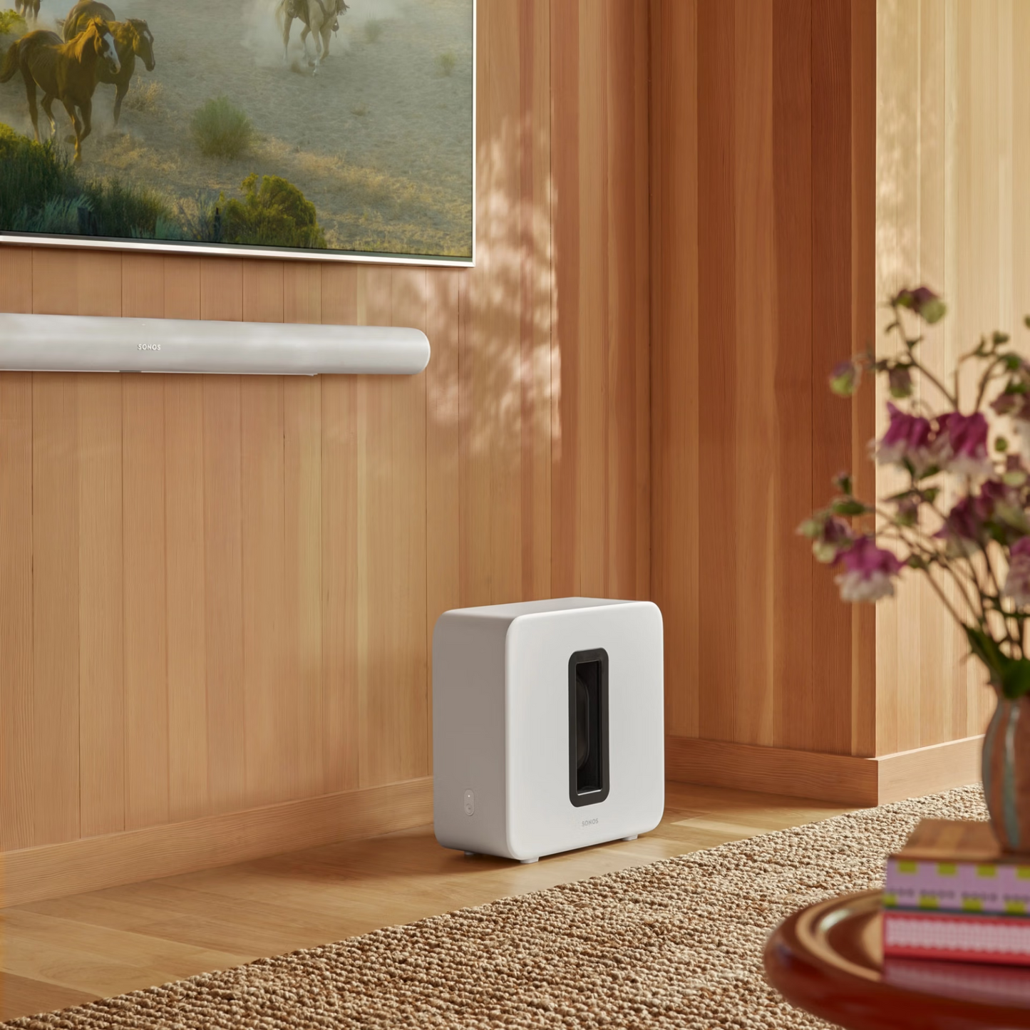 In einem modernen Wohnzimmer mit holzgetäfelten Wänden stehen ein montierter Fernseher, ein Sonos Sub 4 Subwoofer, der für seinen dynamischen Klang und Force-Cancelling-Effekt bekannt ist, eine weiße Soundbar darunter und ein Tisch mit Blumen im Vordergrund.