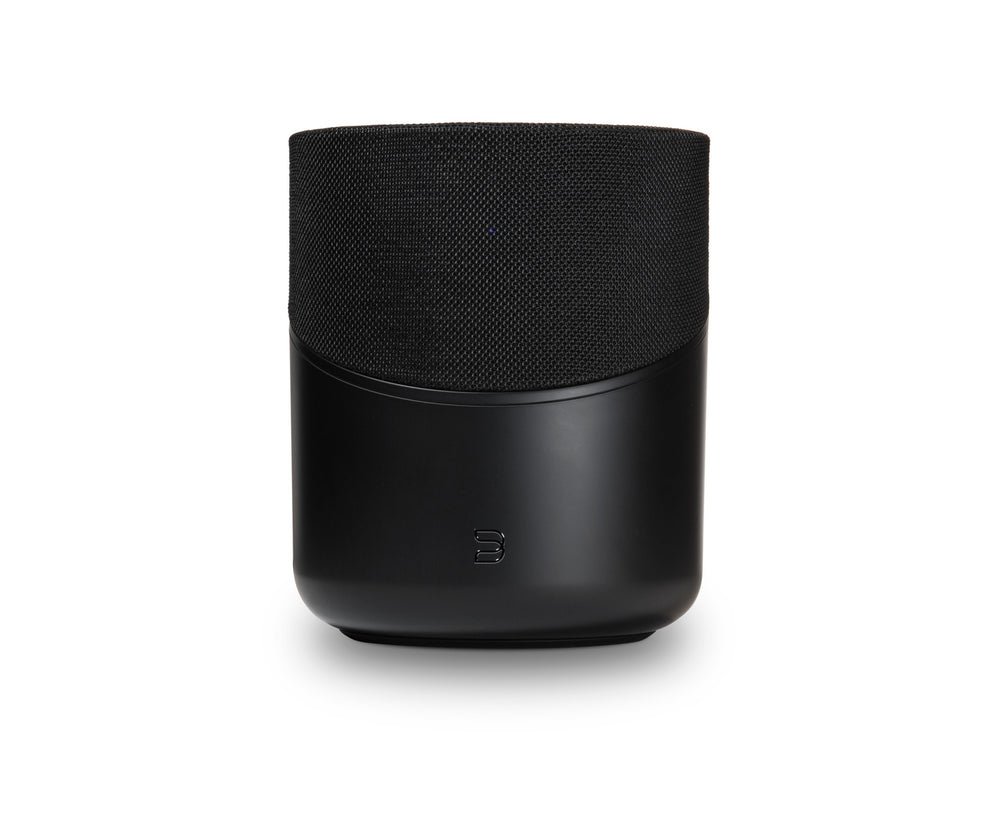 Der Bluesound PULSE M in Schwarz hat eine stoffbespannte Oberseite, eine glatte Unterseite und ein kleines Frontlogo. Dieser kabellose Lautsprecher bietet HiFi-Stereoklang und 360° Klangverteilung. Abgebildet vor einem weißen Hintergrund.