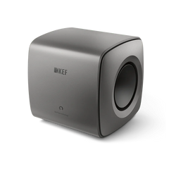 Der KEF KC62 Subwoofer in Titanium Grey besticht durch sein minimalistisches Design mit abgerundeten Kanten, einem Logo auf der Vorderseite und einem runden Seitenlautsprecher.
