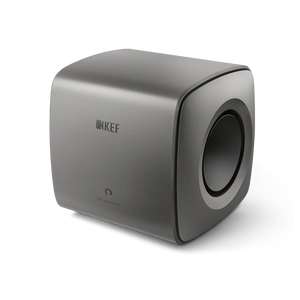 Der KEF KC62 Subwoofer in Titanium Grey besticht durch sein minimalistisches Design mit abgerundeten Kanten, einem Logo auf der Vorderseite und einem runden Seitenlautsprecher.