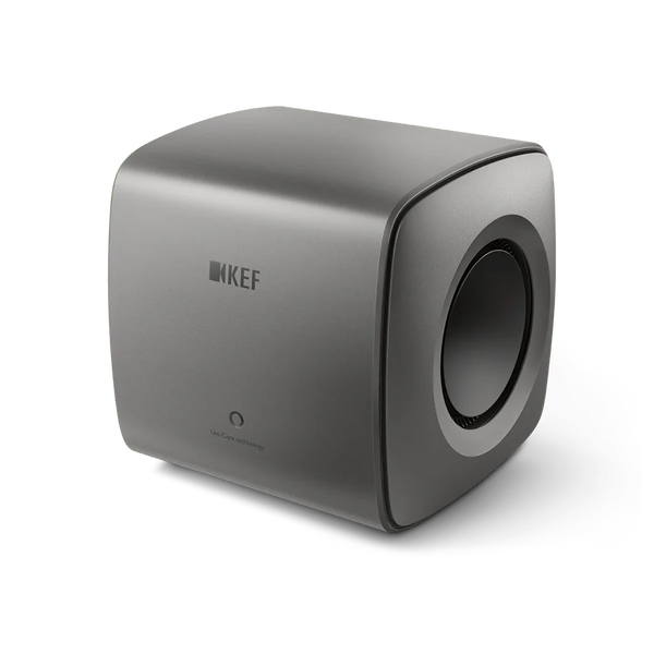 Der KEF KC62 Subwoofer in Titanium Grey besticht durch sein minimalistisches Design mit abgerundeten Kanten, einem Logo auf der Vorderseite und einem runden Seitenlautsprecher.