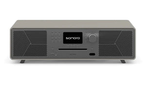Ein Sonoro Meisterstück (Gen.2) in Sandstein (matt) - Satingrau mit zentralem Digitaldisplay, Bedientasten, Lautstärkeregler und zwei Lautsprechergittern, mit Dynamic Bass Funktion für verbesserte Audioqualität auf weißem Hintergrund.