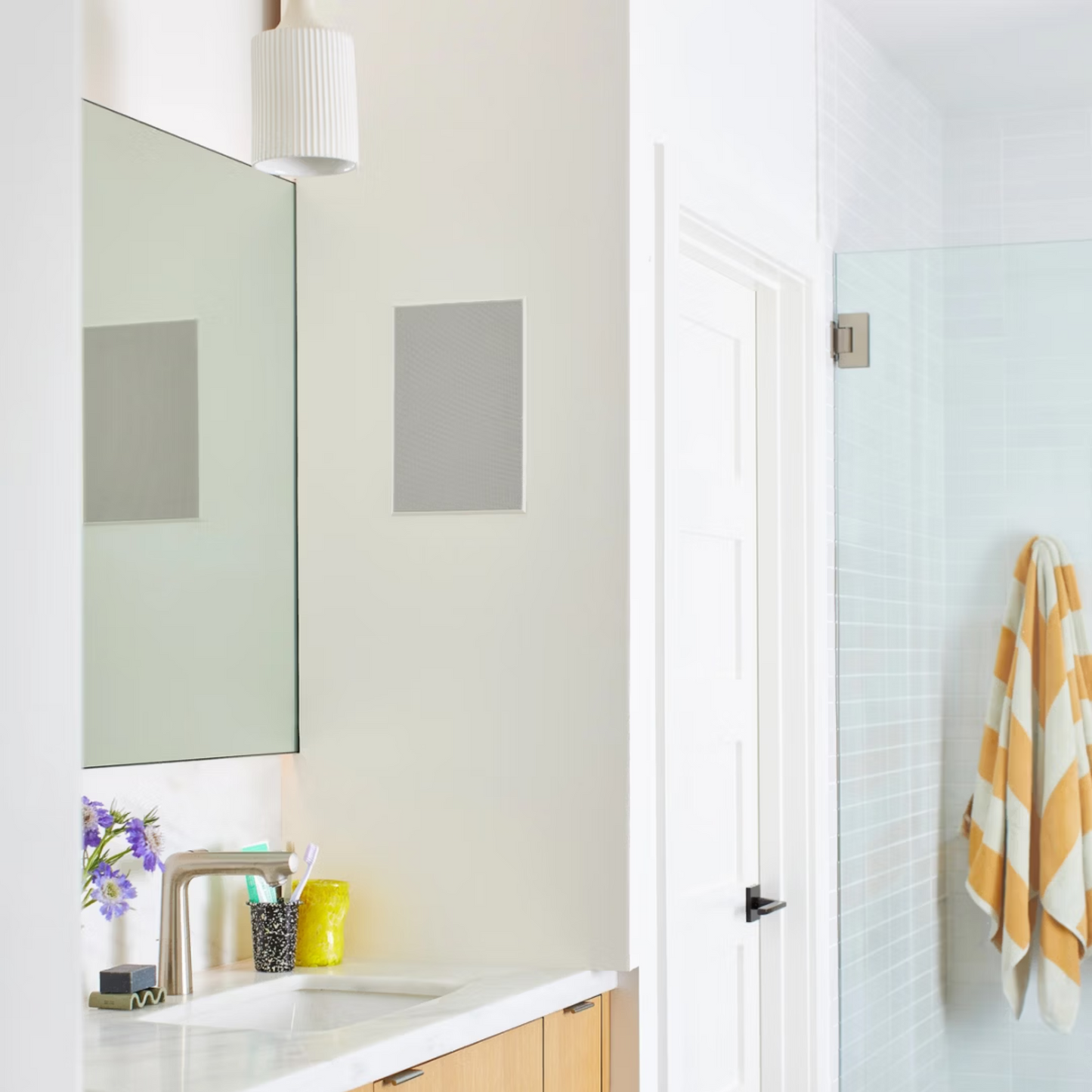 Ein modernes Badezimmer mit weißer Arbeitsplatte, Holzschränken, Spiegel, lila Blumen, Toilettenartikeln und einer Dusche mit Glastür und gelb gestreiftem Handtuch - ideal zum Entspannen mit dem immersiven Soundreference Sonos In-Wall Speaker von Sonos und Sonance Paar.
