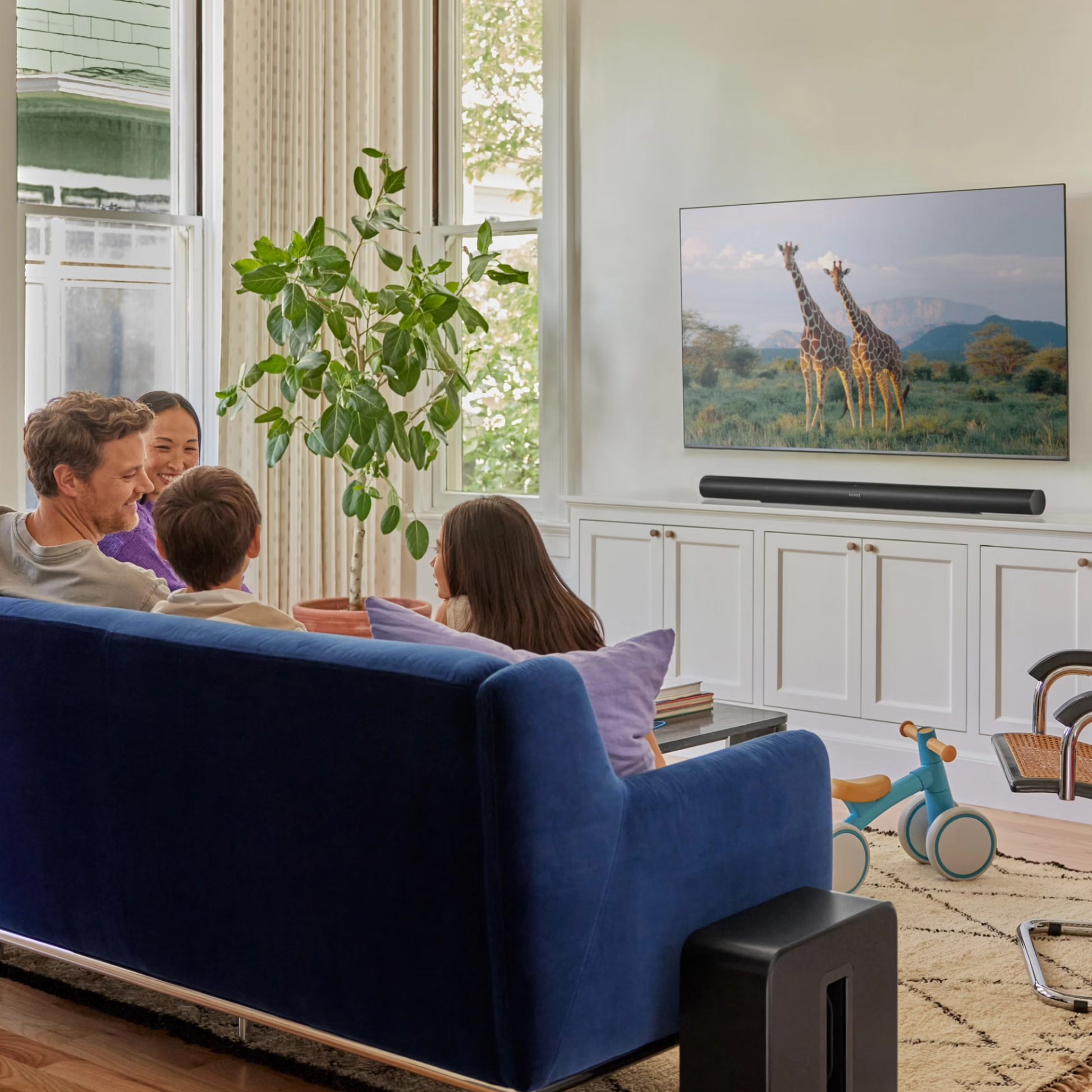 Eine Familie entspannt sich auf einem blauen Sofa in einem hellen Wohnzimmer und schaut Fernsehen mit kristallklarem Dialoge und beeindruckendem Dolby Atmos Sound von der Sonos Arc Ultra Soundbar. Spielzeug und eine Pflanze sorgen für Gemütlichkeit, während Giraffen auf dem Bildschirm erscheinen.