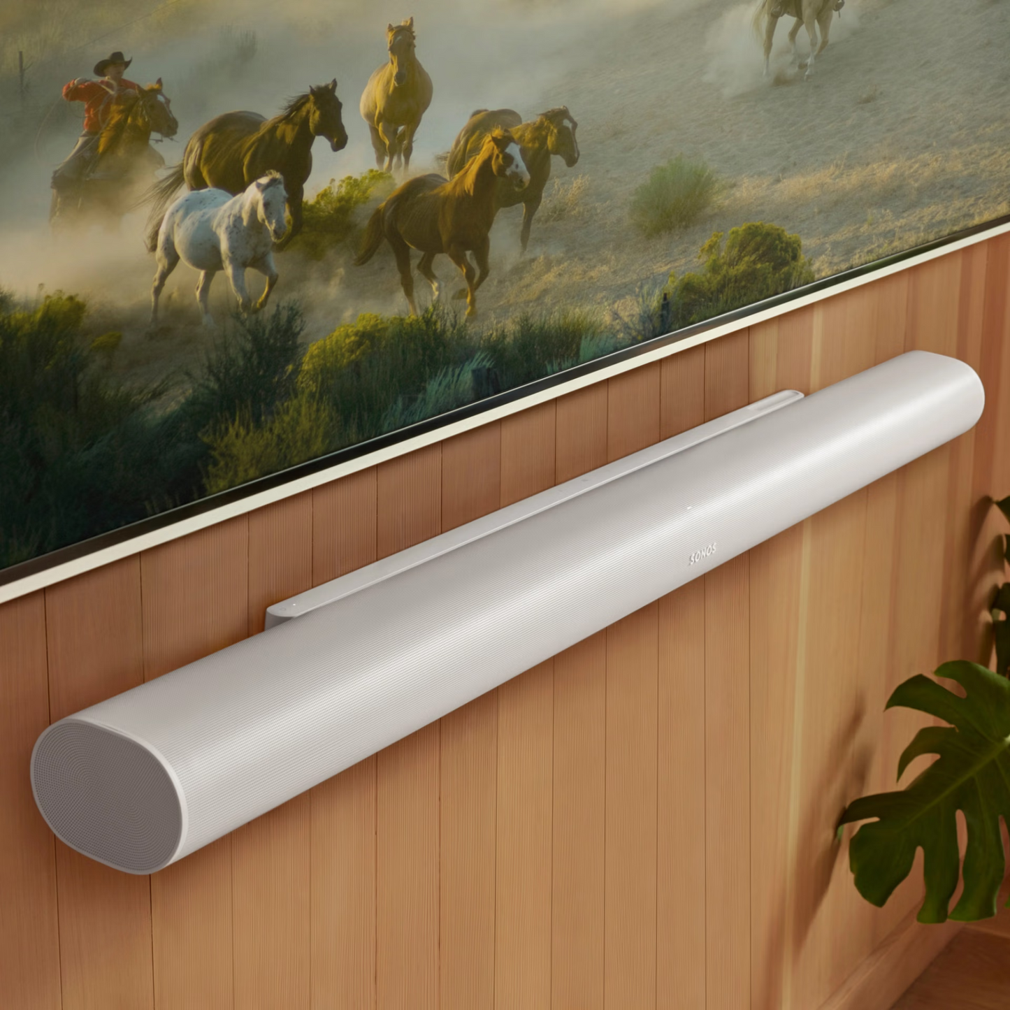 Die Sonos Arc Ultra Soundbar, ein elegantes weißes Modell mit Dolby Atmos, ist an einer holzgetäfelten Wand unter einem Fernseher angebracht, der laufende Pferde zeigt. In der unteren rechten Ecke erscheint ein grünes Pflanzenblatt.