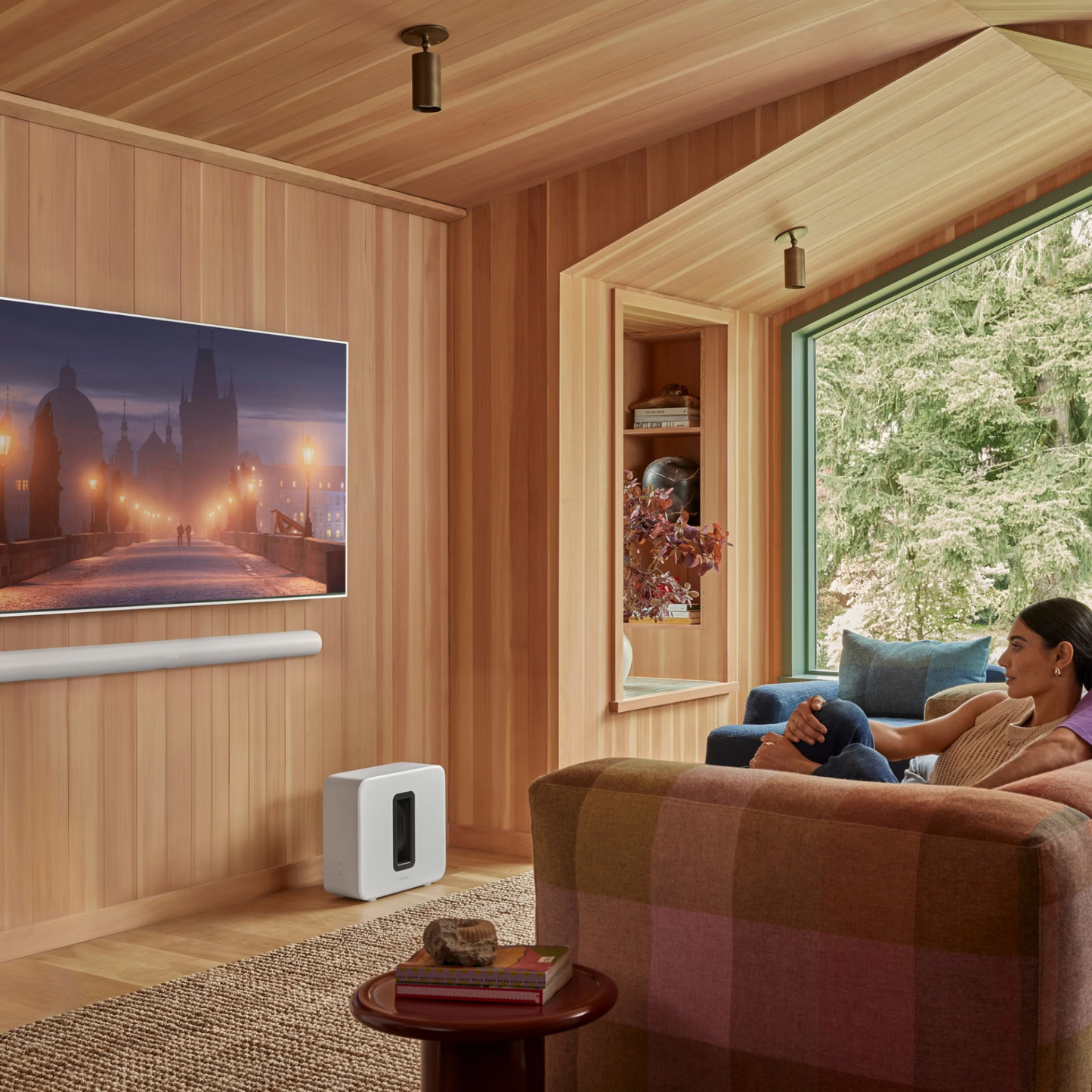 Eine Frau entspannt sich auf einem karierten Sofa in einem holzgetäfelten Wohnzimmer. Unter dem Fernseher liefert die Sonos Arc Ultra Soundbar mit Dolby Atmos kristallklare Dialoge und immersiven Klang im ganzen Raum.