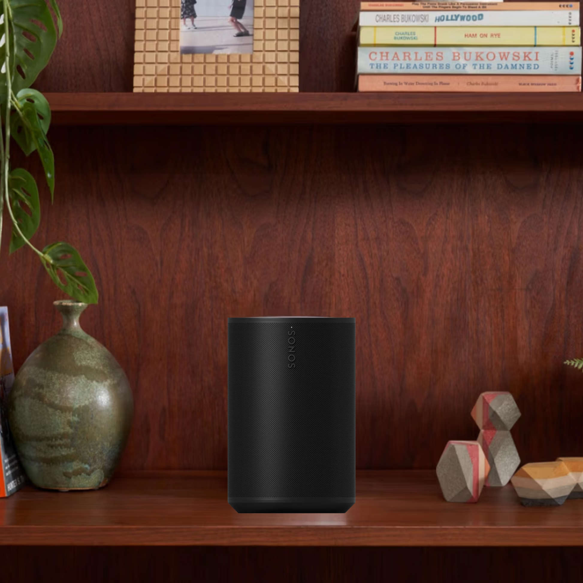 Ein schwarzer Sonos Era 100 Smart Speaker steht auf einem Holzregal zwischen einer grünen Keramikvase mit einer Pflanze und geometrischem Dekor, während auf dem oberen Regal dahinter Bücher und ein gerahmtes Foto zu sehen sind.
