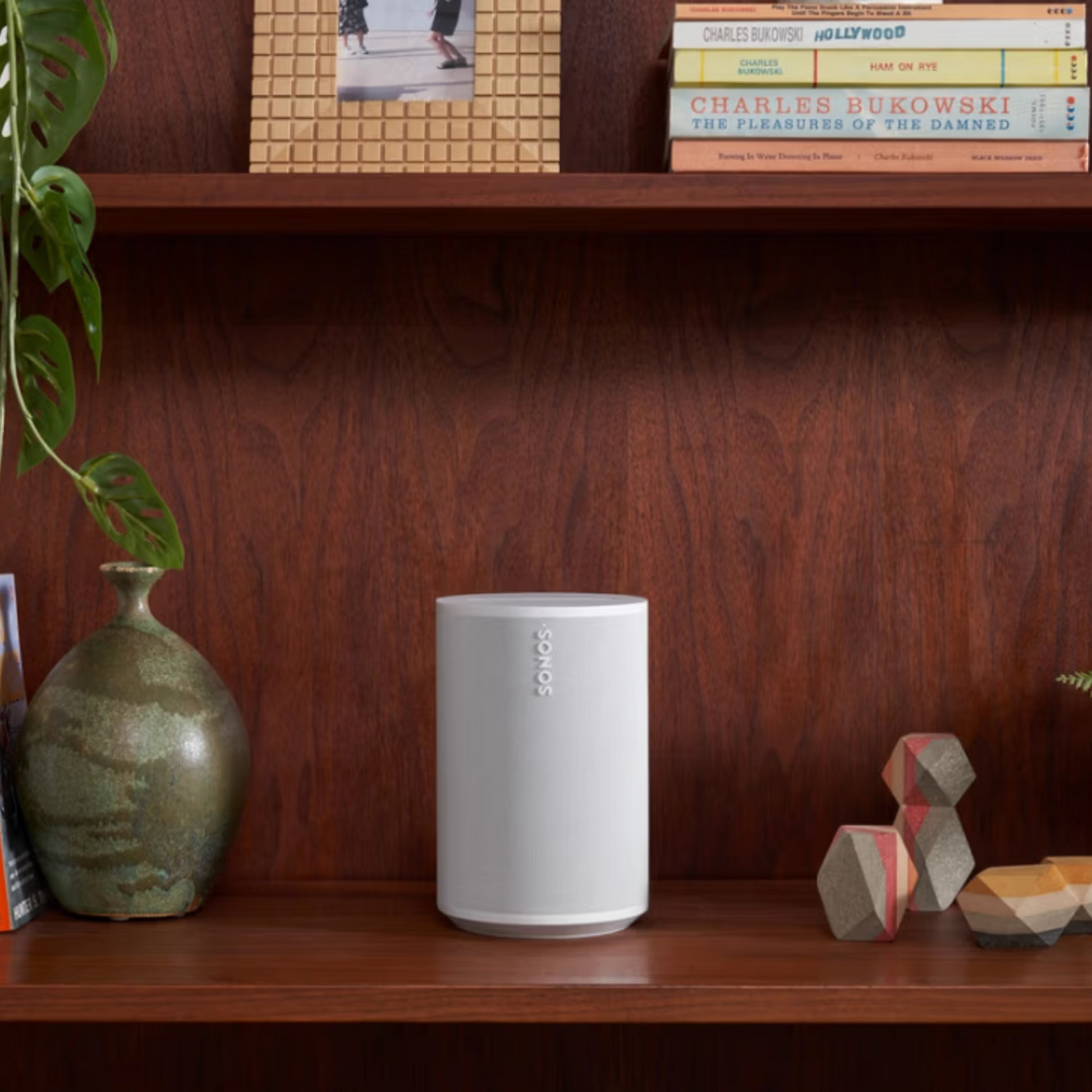 Ein weißer Sonos Era 100 Smart Speaker steht auf einem Holzregal, zwischen einer grünen Keramikvase mit einer Pflanze auf der linken Seite und geometrischem Dekor auf der rechten Seite. Auf dem Regal darüber sind Bücher und ein gerahmtes Foto zu sehen.
