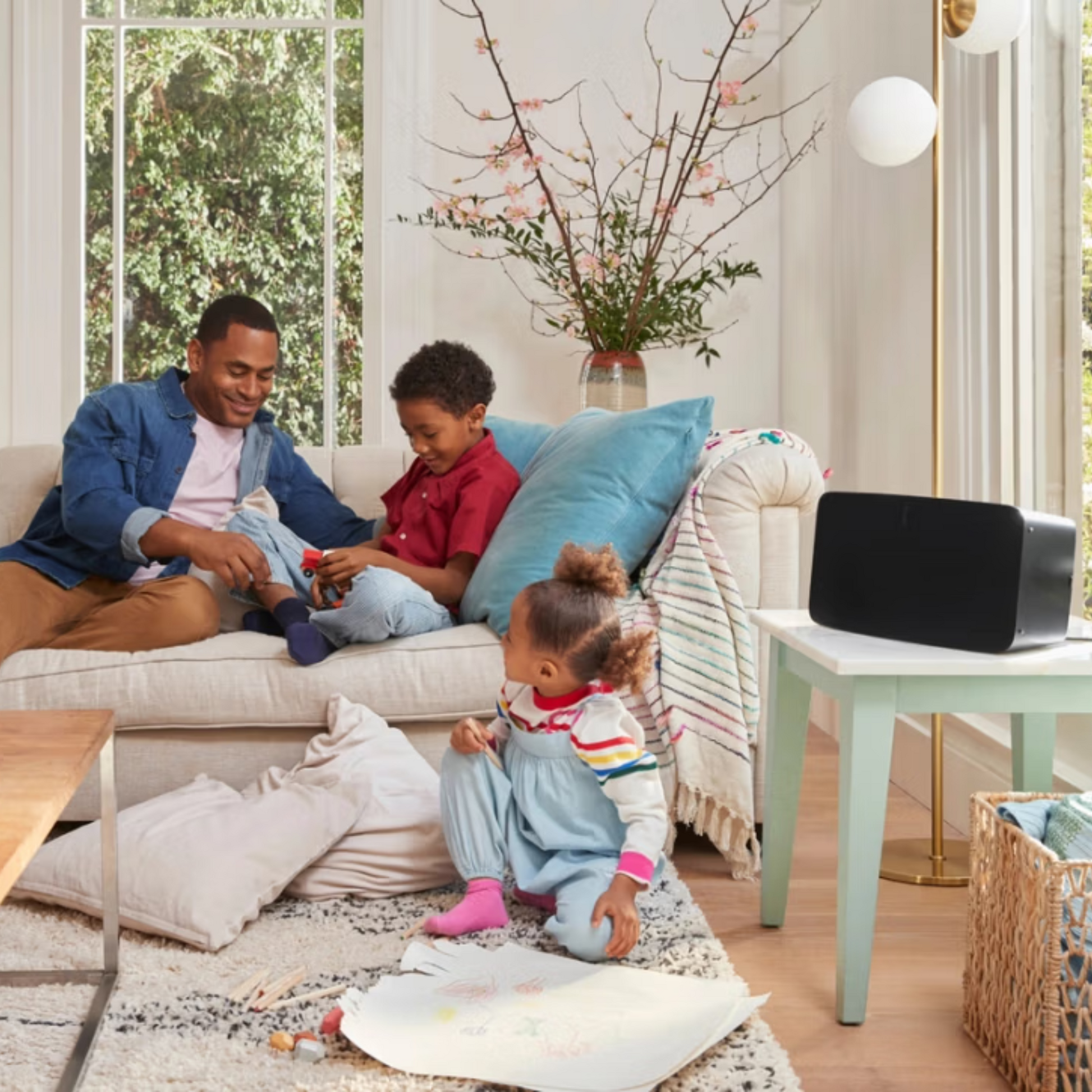 Ein Mann und zwei Kinder genießen in einem sonnendurchfluteten Wohnzimmer mit der Sonos-App hochauflösenden Sound aus dem Sonos Five Smart Speaker. Der Mann und ein Kind entspannen sich auf einer weißen Couch, während ein anderes auf dem Teppich in der Nähe der großen Fenster zeichnet.