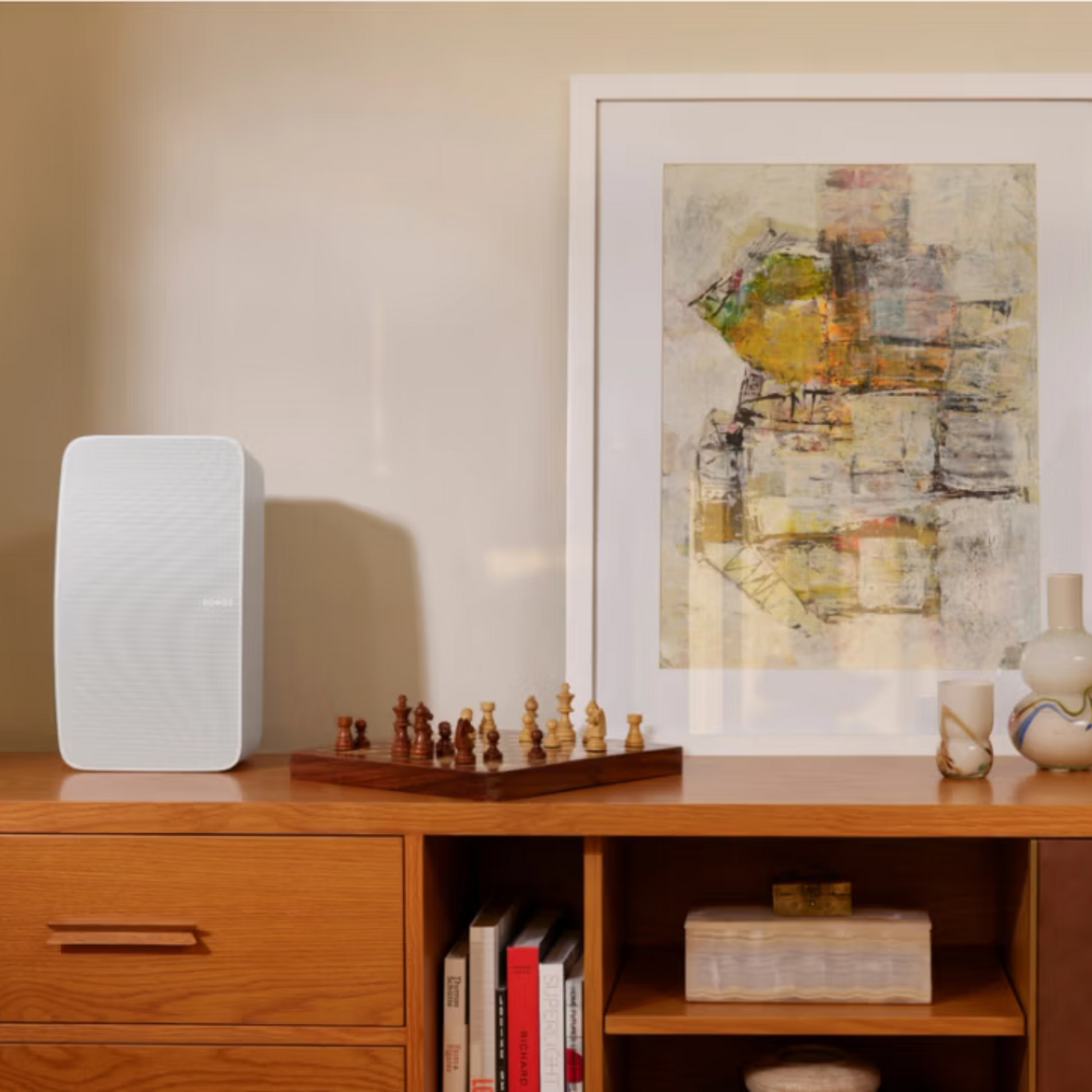 Auf einem minimalistischen Holzsideboard stehen ein weißer Sonos Five Smart Speaker (hochauflösendes Audio), ein Schachbrett mit Figuren, Vasen und Bücher. Darüber hängt ein gerahmtes abstraktes Gemälde an der beigen Wand.