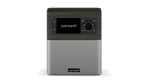 Das Sonoro Stream in Mooreiche (dekor) - Silber ist ein rechteckiges DAB+-Digitalradio mit Bedientasten und einem schwarzen Display auf der Oberseite sowie einem silbernen Lautsprechergitter darunter. Das Sonoro-Logo erscheint sowohl auf dem Bildschirm als auch auf dem Gitter.