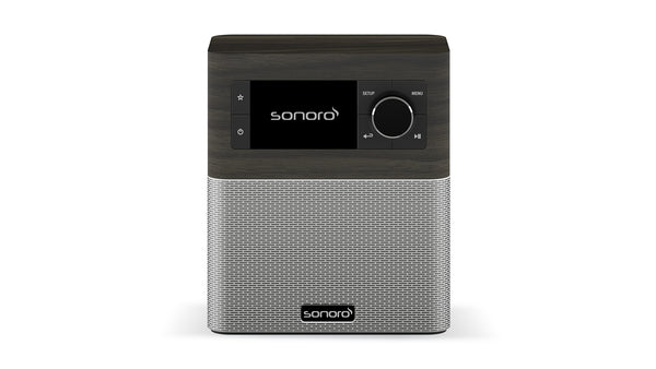 Das Sonoro Stream in Mooreiche (dekor) - Silber ist ein rechteckiges DAB+-Digitalradio mit Bedientasten und einem schwarzen Display auf der Oberseite sowie einem silbernen Lautsprechergitter darunter. Das Sonoro-Logo erscheint sowohl auf dem Bildschirm als auch auf dem Gitter.
